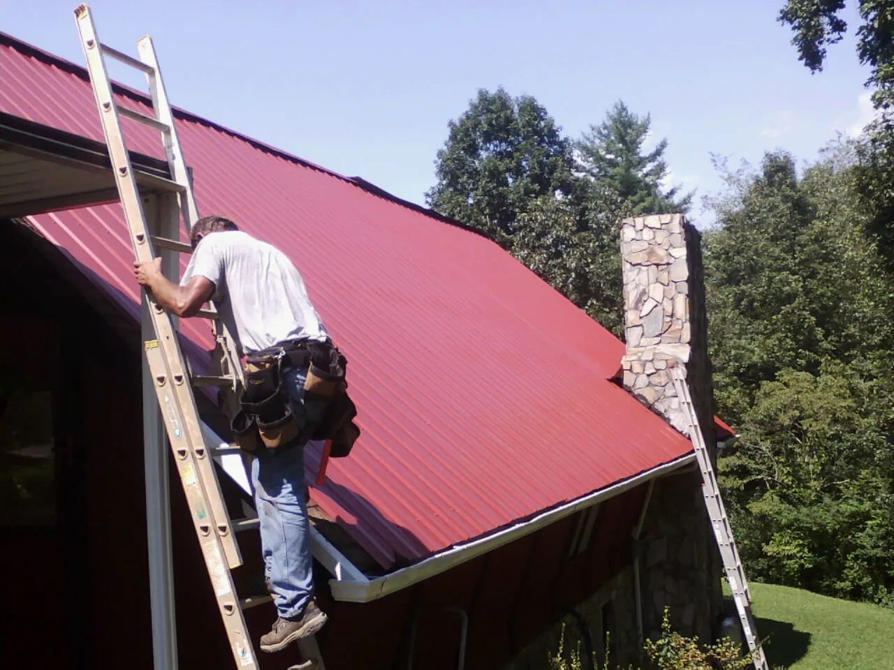 Roofer on a ladder beside a red metal roof and stone chimney — Soffit & Fascia Repair by Roofing Brothers in Bonners Ferry